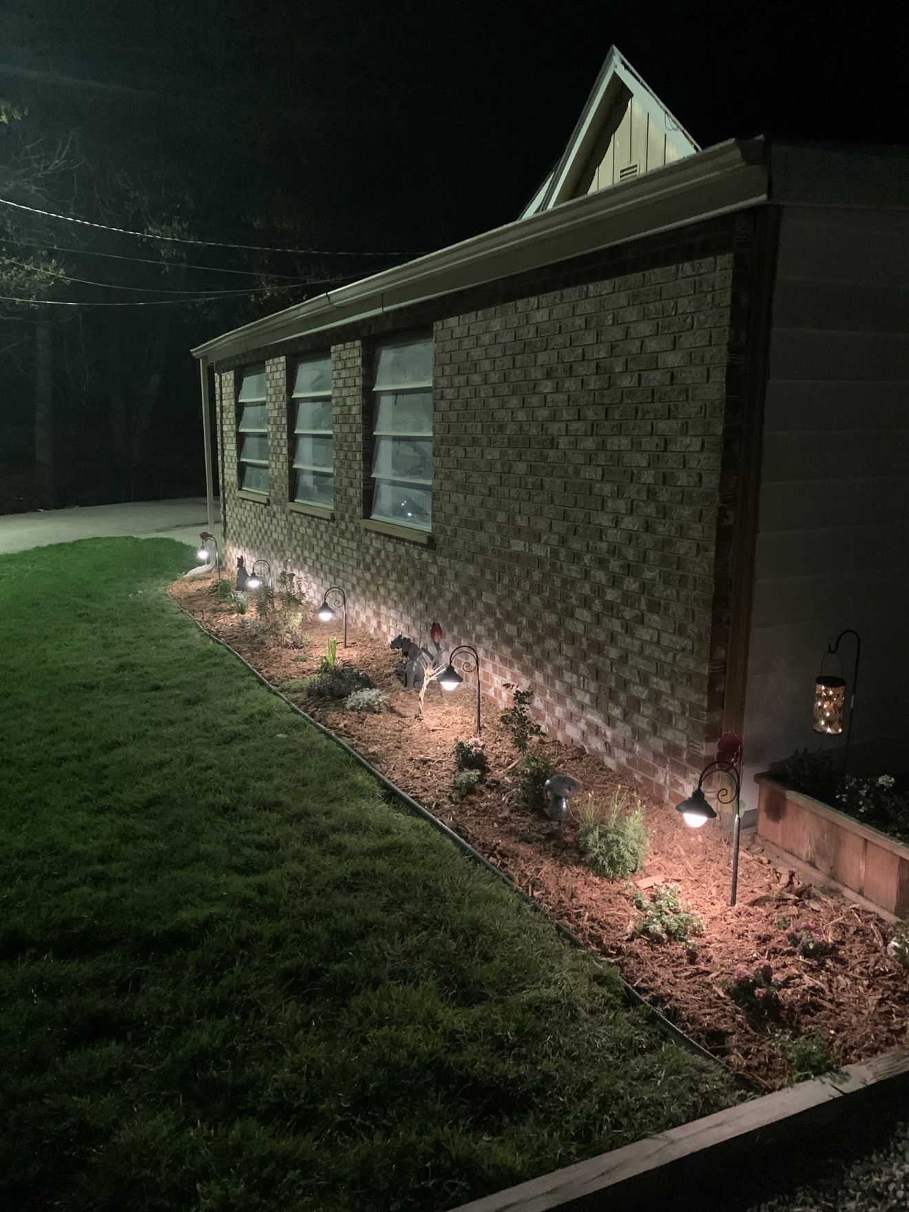 Mulched landscape bed along a brick wall lit by shepherd's-hook path lights at night